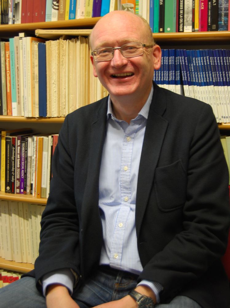 Torso portrait of Mark Pearce sitting in front of a bookshelf smiling at the camera