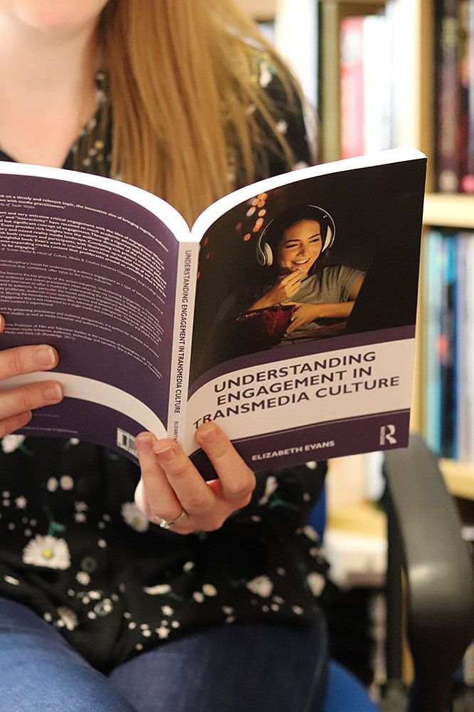 A photograph of a woman sitting down and reading a book entitled 'Understanding Engagement in Transmedia Culture'.