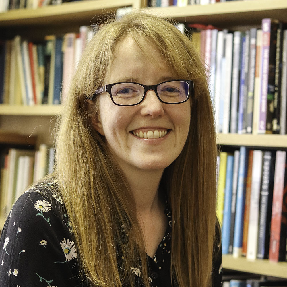 A headshot of Liz Evans standing in front of a bookcase and smiling at the camera.