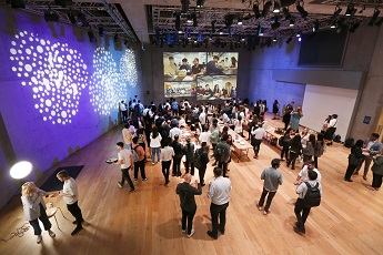 A photo of an event indoors with lots of people in a room with large projected images on the wall. The photo is taken from a high up angle, looking down across the room.
