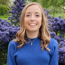 Photo of Fiona Frost, doctoral research student, stood outside with purple flowers and trees in the background. Fiona is smiling and wearing a blue long sleeve jumper and a necklace.