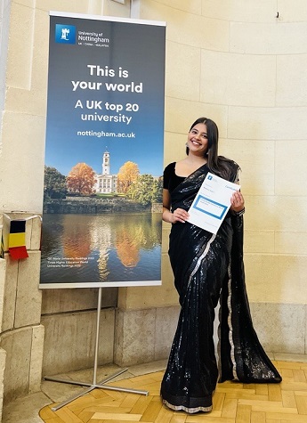Mahima Udani wearing a long black gown stood by a Nottingham University banner that reads "This is your world. A UK top 20 university."