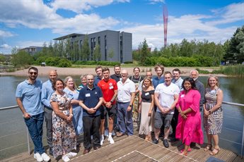 19 male and female participants from cohort 13 of the Help to Grow: Management programme stand on decking in front of the Jubilee campus lake.