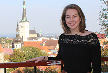 Kathryn Moore smiles at the camera from a balcony in Estonia, overlooking a medieval town including church spire