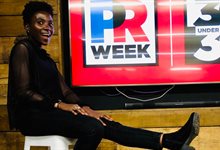 Tobi-Ruth Adebekun sitting smiling in front of a 'PR week 30 under 30' sign on a television screen