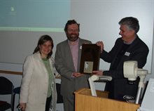 A woman and two men standing behind a lectern with a whiteboard behind smile at the camera, while the man in the middle is presented with an award