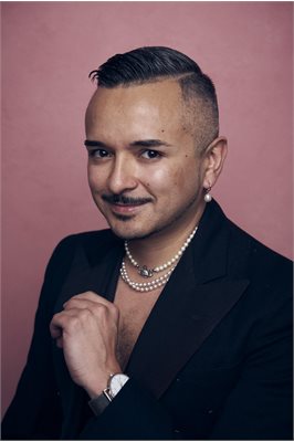 A portrait shot of David Martinez who is sitting against a dusk pink background. He is a brown, Latin man with a moustache and an undercut haircut. He is wearing two pearl necklaces and a dark navy suit.and