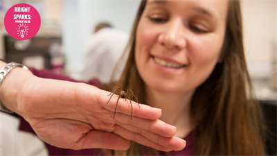 Close up image of a white, smiling feamle holding a spider on her hand
