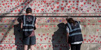 A wall covered in small painted red hearts, two people with their backs to the camera are adding hearts