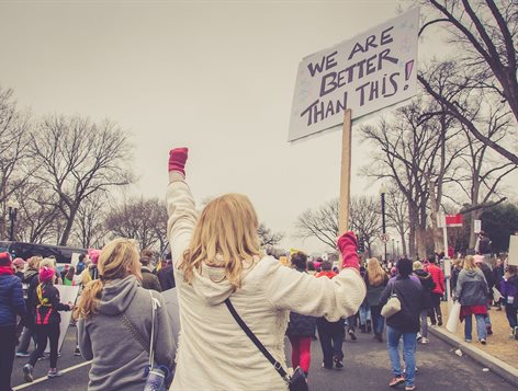 The back of a group of people during a demonstration. Focal point is a person raising their fist holding a sign which reads "we are better than this"