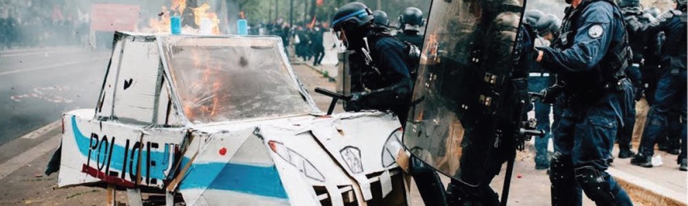 Riot police destroying a cardboard police car  which is on fire