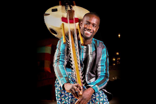 A photo of Sura Susso in native Gambian clothing smiling, with his kora resting on his right shoulder