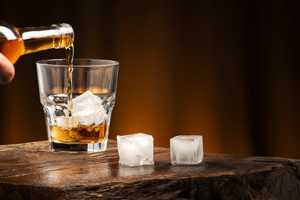 Pouring whiskey into a glass with ice. A glass of alcoholic beverage on a wooden table.
