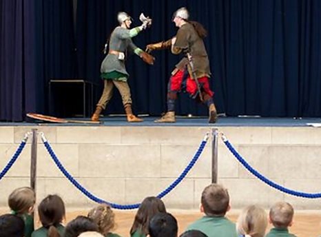 Photograph of two figures in Viking clothes fighting on stage in front of an audience of schoolchildren.