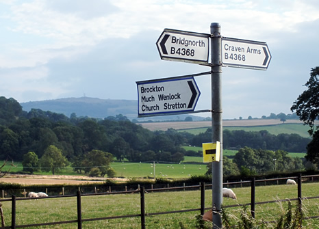 English road sign with three signs pointing in different directions and blue skies and green fields behind.