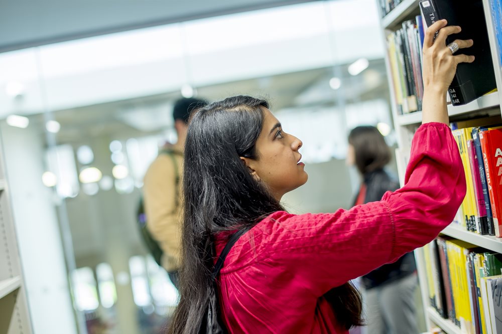 South Asian women with long black hair and wearing a red top reaching up to take down a book from a library shelf