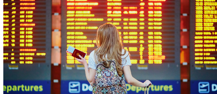 Photograph of a person holding their passport stood in front of an arrivals/departures board