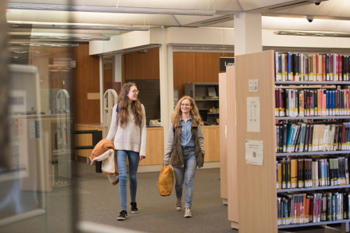 Two smiling students walking beside library bookshelves