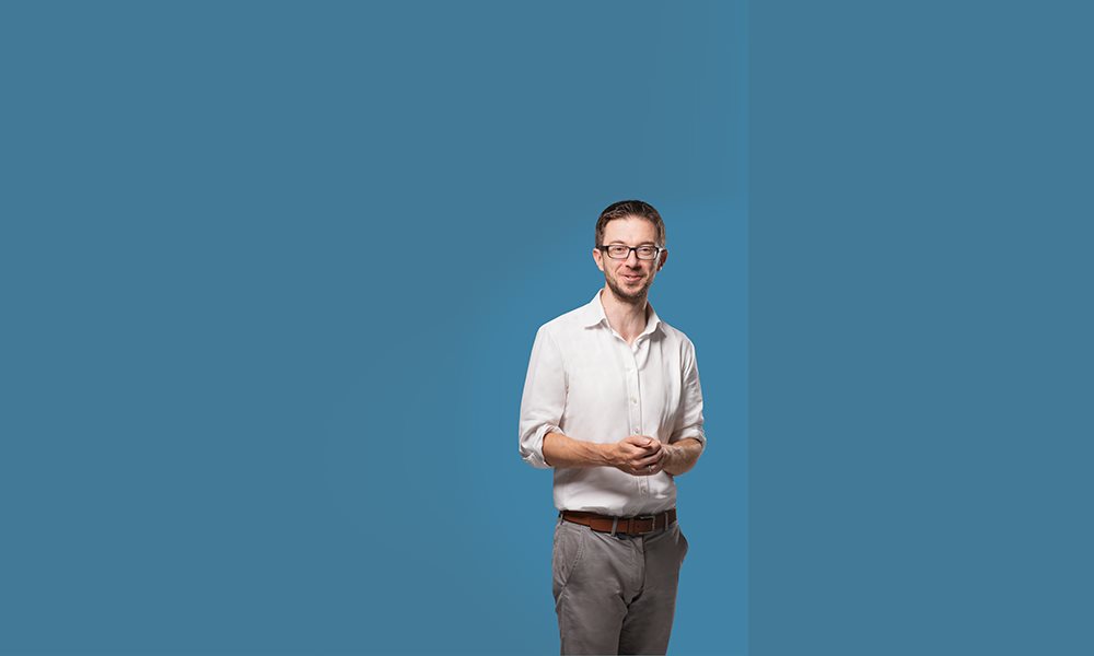 A white man with a bear and glasses, wearing a white shirt and grey trousers, stands in front of a blank blue background