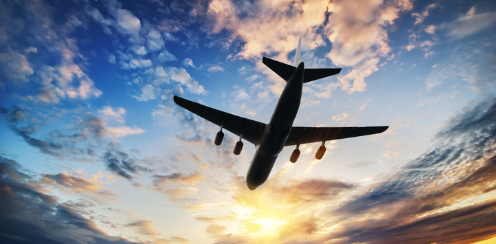 An aeroplane in flight, seen from below in partial silhouette, against a blue sky with white clouds and bright sunlight