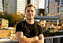 A profile picture of Freddie who wears a black t-shirt and smiles against a cityscape