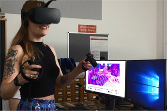 Young white woman (Aja Ireland) wearing a VR headset and holding controllers looking to the right of the camera with several large monitors behind her