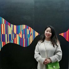 Young Asian woman in a long white jumper stood in front of a wall decorated with abstract artwork made with colourful lines