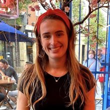 Head and shoulders photograph of a young smiling white woman with long brown hair wearing a black top standing outside in front of a busy cafe