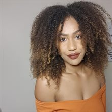 Head and shoulders photograph of a young black woman with curly light brown hair wearing an off-shoulder orange top in front of a plain grey background.