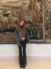 A photo of Lucy Noble standing in front of a tapestery