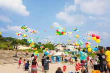 A large group of children and adults standing on a beach releasing balloons into the sky