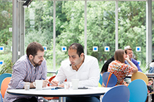 Two male students sitting at a round table having a discussion, with other students in the background