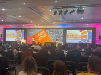A group of students sitting down facing a stage with large bright slides and a speaker addressing the audience for the Enactus Nottingham at the National Expo 2025