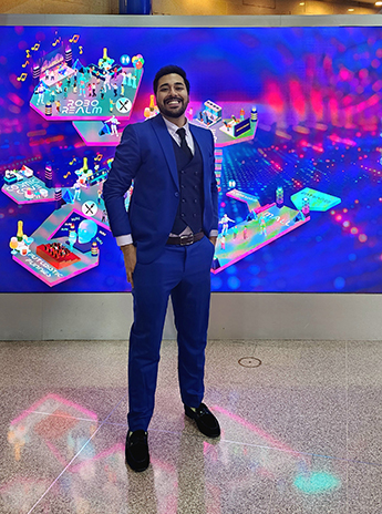 Full length photo of Saad Sher Dil Khan stood in front of a vibrant colourful digital screen showing various graphics on a bright blue background. He is wearing a blue three piece suit with his hands in his pockets, smiling at the camera.