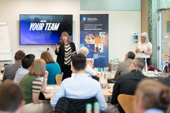 Participants of cohort 2 of the EPSTL programme sit in groups around tables. They listen to Dr Julie Blant as she delivers a Spotlight Profiling workshop. A screen showing the words 'Your team' is visible behind her and Dr Hannah Noke stands off to one si