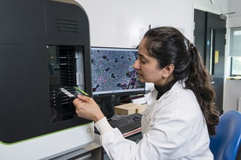 Sunny Sunshine sits at a desk within her lab, holding a sample on a slide next to a machine. She wears a white lab coat and a computer screen with an image of an enlarged sample is visible behind her.