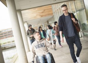 Two students in the foreground , one  in a wheelchair, chatting whilst travelling along the lakeside walkway on Jubilee Campus. Other students can be seen in the background.