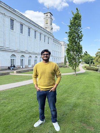 Aakash Parameswaran with black short curly hair wearing a bright yellow jumper, grey trousers and white trainers standing on grass in front of white Trent Buiding at the University Park Campus in Nottingham
