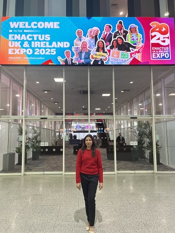 Aiswarya Krishnan with long black hair, wearing a red jumper and black trousers standing in front of large glass doors below a bright Enactus Expo 2025sign
