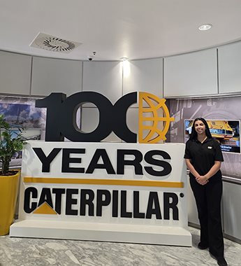Undergraduate student Anika Dhunna standing next to a large display celebrating the manufacturing company Caterpillar Inc.&rsquo;s centennial. The sign features a bold black &lsquo;100&rsquo; with the zero on the right designed as a yellow globe symbol. Below it, the words