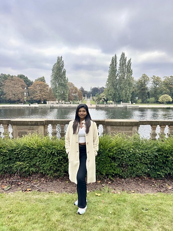 Clara Benita Jesula Justin Thomas with long black hair wearing a long cream coat and white top, black trousers and white trainers standing on grass in front of the lake at the University Park Campus in Nottingham