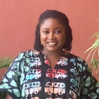 Janet Torhee smiling, with short black curly hair wearing a bright batik printed top, standing in front of a terracotta wall