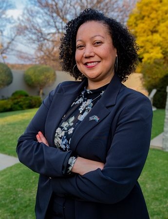 Photo of MBA graduate Mecia Peterson. Mecia is stood outside with trees visible in the background. Mecia is wearing a dark navy jacket and a floral top and is folding her arms across her body.