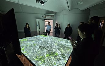Participants from Pecege visit the City as Lab installation during their programme. They are shown standing around the model, a lit up digital projection of the city of Nottingham on a textured table, as a member of the City as Lab team talks them through
