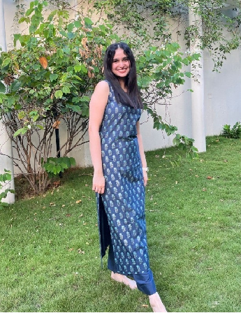 Serah Dominic standing in front of some tall bushes on grass, wearing a dark blue printed dress, has long dark hair and smiling.
