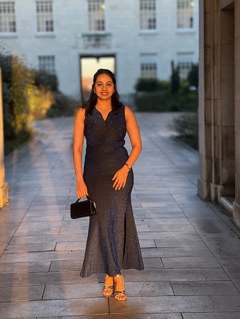 Shreya Dathathraya with long black wavy hair smiling, wearing a black glitter dress and silver shoes, standing in front of the white brick Trent Building at the University Park campus