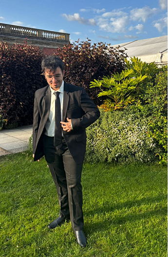 Shubham Thakkar smiles, with short black hair and glasses, wearing a black suit and tie over a white shirt, standing on grass in front of bushes.