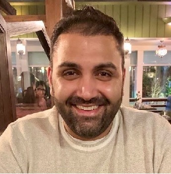 Sunny Sahdev with black short hair and beard and smiling wearing a cream jumper