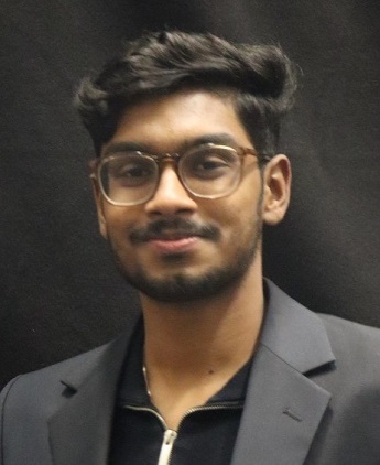 Thejeshwar Karthikeyan with short dark hair and beard, wearing glasses and smiling. Wearing a dark grey blazer and black zip top