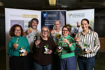 Tom Hignett and his presentation group pose for a photo after winning the award for best presentation. The group of 6 people all smile, each holding a celebratory drink in one hand and their prizes, stuffed toy ducks, in the other.